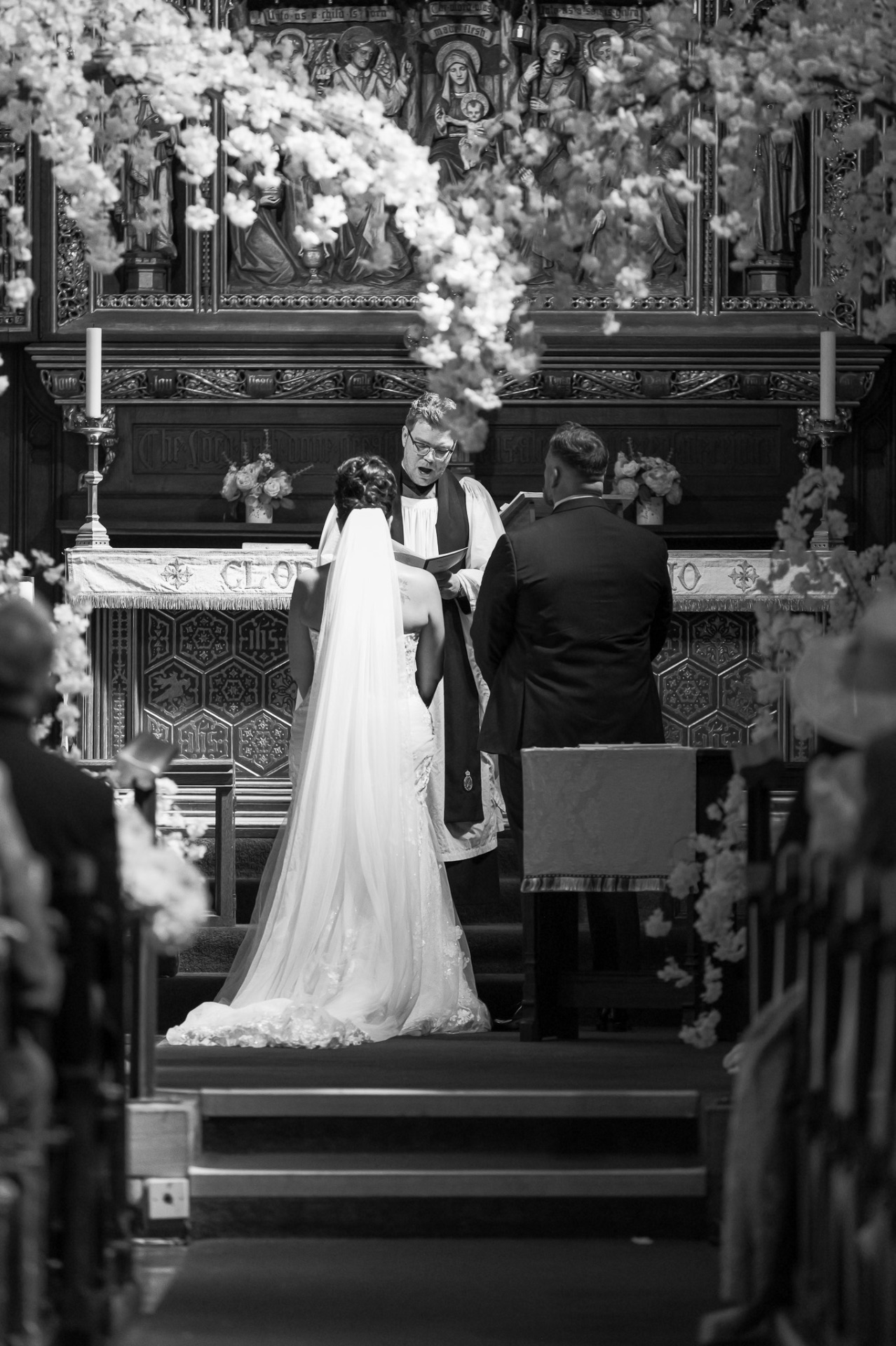 Black and white photograph of bride and groom exchanging vows at a flower-adorned church altar, wedding photography Worcestershire & Midlands